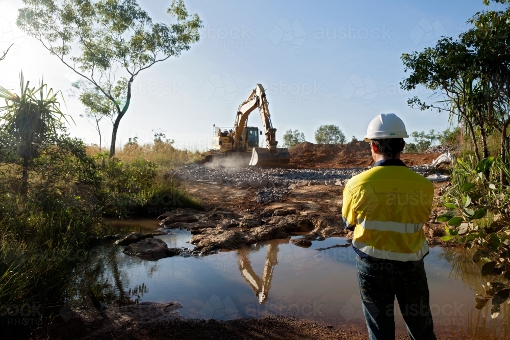 Industrial worker overseeing a large digger moving rocks on an industrial building site - Australian Stock Image