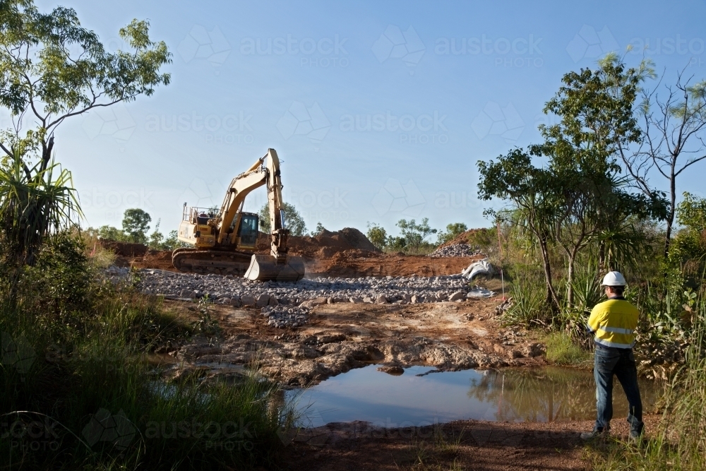 Industrial worker overseeing a large digger moving rocks on an industrial building site - Australian Stock Image