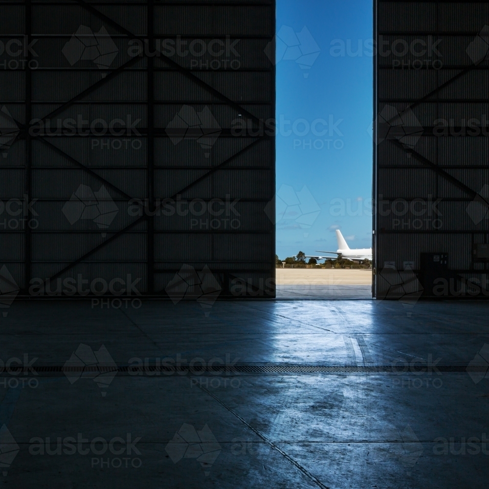 Inside an airport hangar looking out to a plane on the runway through a narrow opening - Australian Stock Image