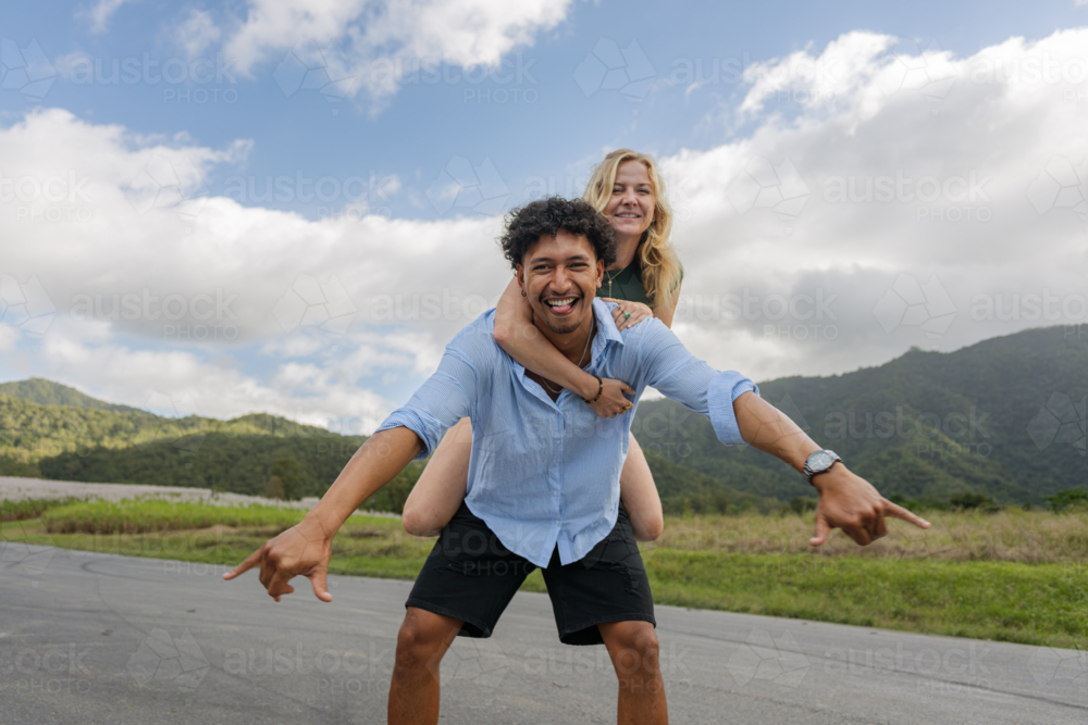 Interracial couple laughing during piggyback ride on a country road - Australian Stock Image