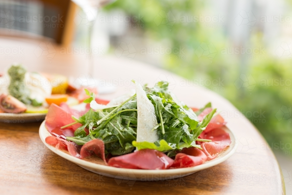 Italian bresaola, rocket and parmesan salad with a glass of wine and tomato salad in the background - Australian Stock Image