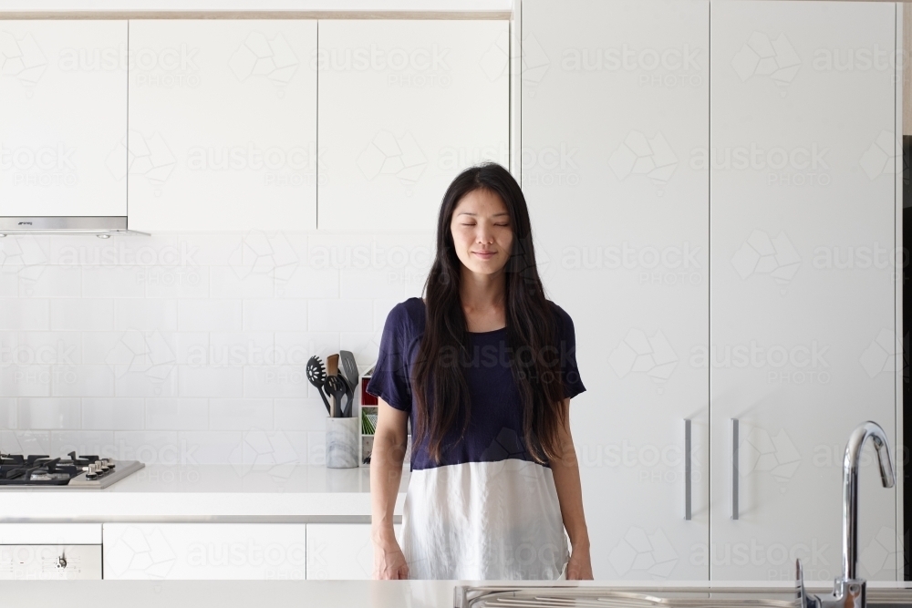 Japanese woman standing in kitchen with eyes closed - Australian Stock Image