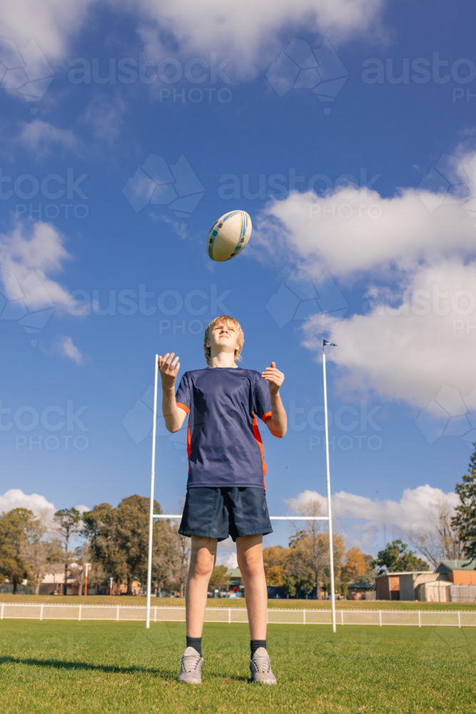 Junior rugby union player standing on field handling ball with goal posts in background - Australian Stock Image