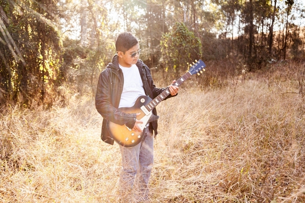 Kid in dark jacket and shades with electric guitar outside - Australian Stock Image