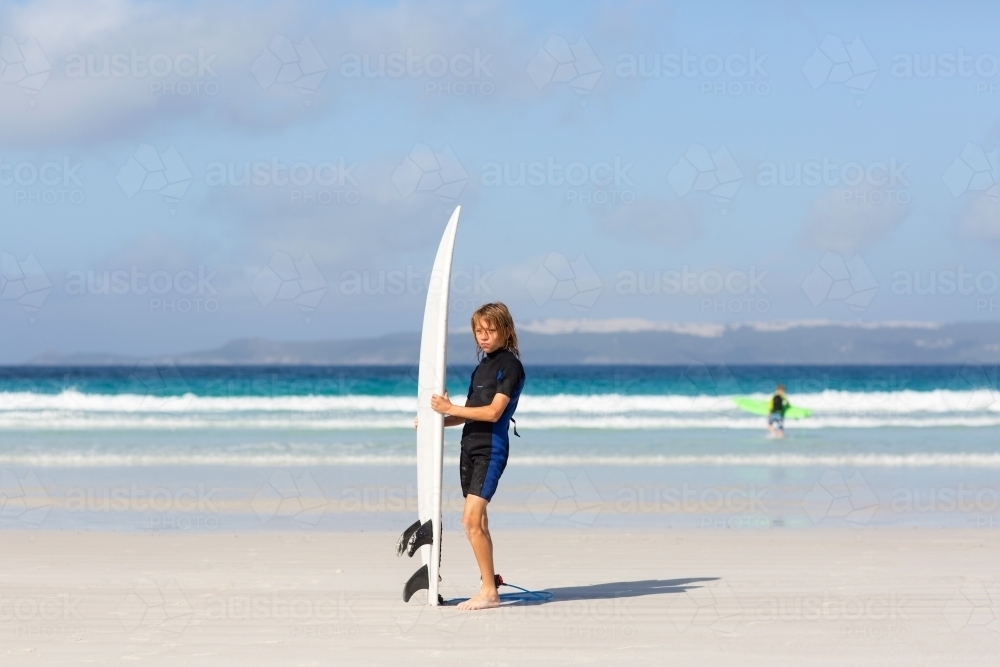 Kid in wetsuit showing off his surfboard on the beach - Australian Stock Image
