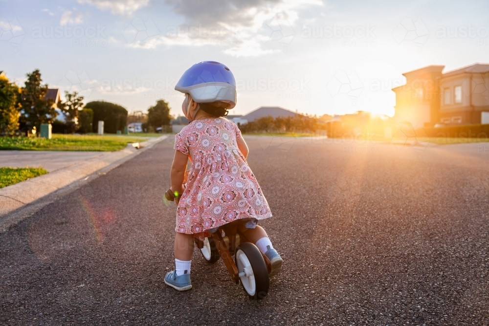 Kid riding balance bike down quiet suburban road wearing helmet in flare of sunset light - Australian Stock Image