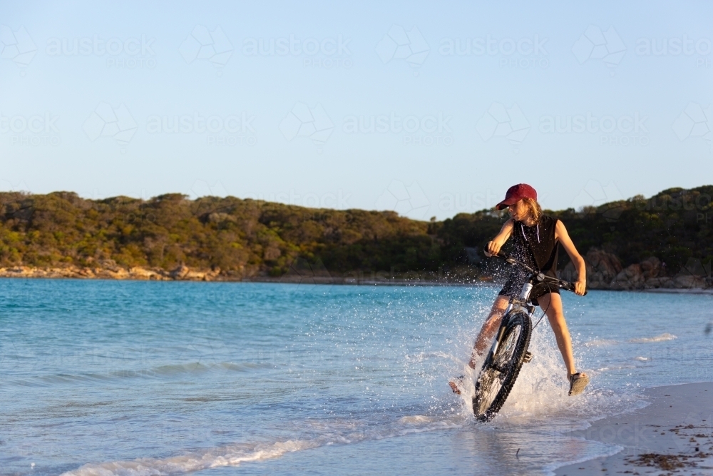 Kid riding bike through water on beach - Australian Stock Image