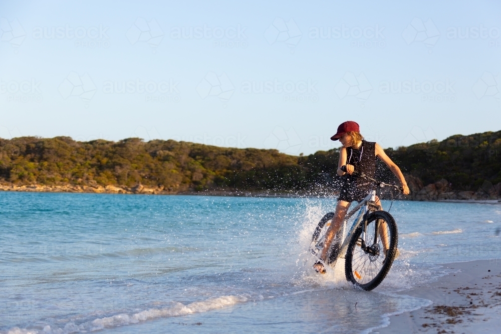 Kid riding bike through water on beach - Australian Stock Image