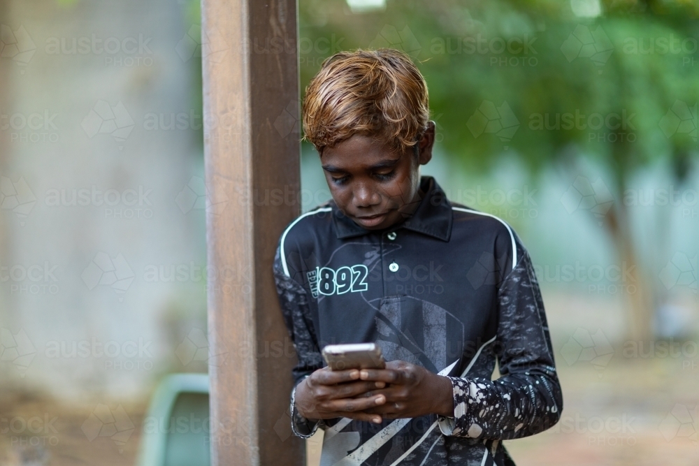 kid with bleached hair looking down at mobile phone while leaning on post - Australian Stock Image