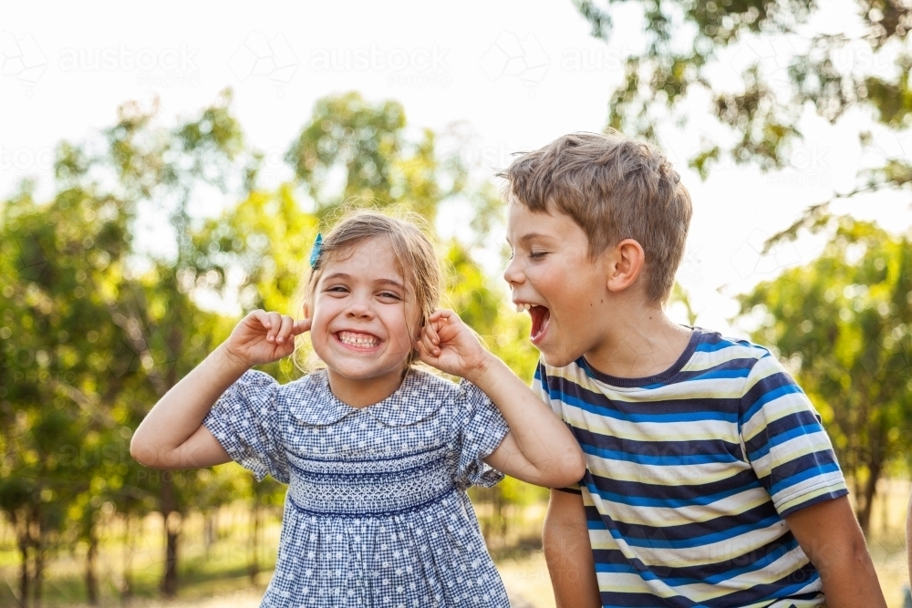 Kids being silly together - boy shouting and roaring at little girl with ears blocked not listening - Australian Stock Image