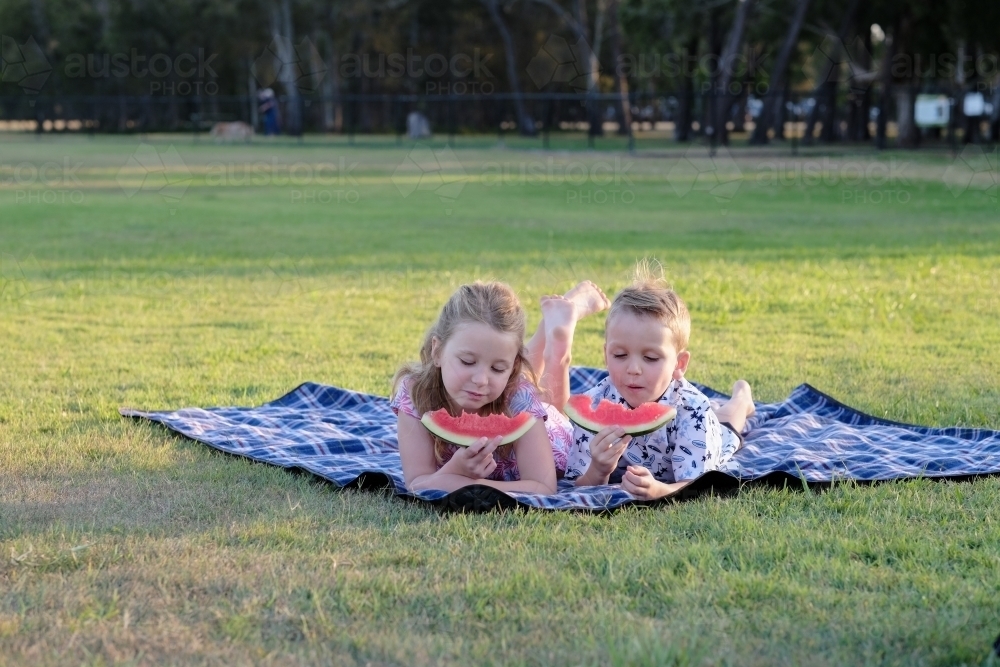 Kids eating watermelon on a picnic blanket in a park - Australian Stock Image