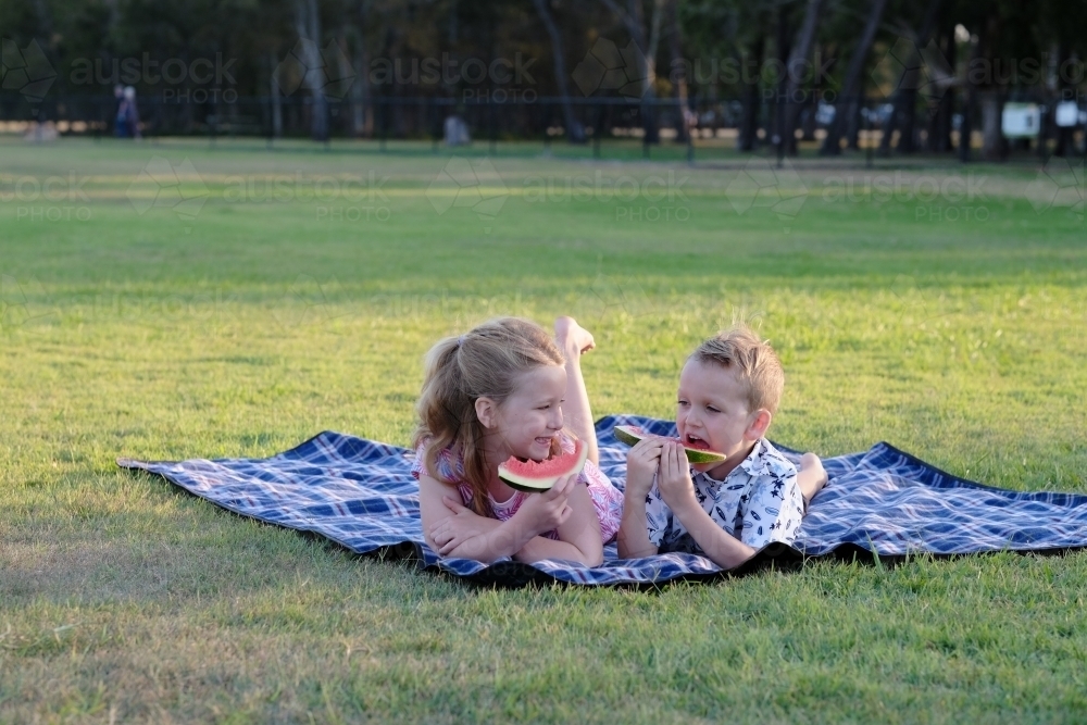 Kids eating watermelon on a picnic blanket in a park - Australian Stock Image