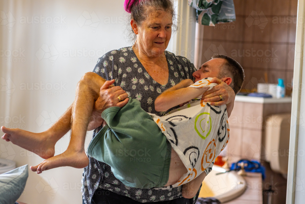 lady carrying her adult son who has cerebral palsy in the house with bathroom through door - Australian Stock Image