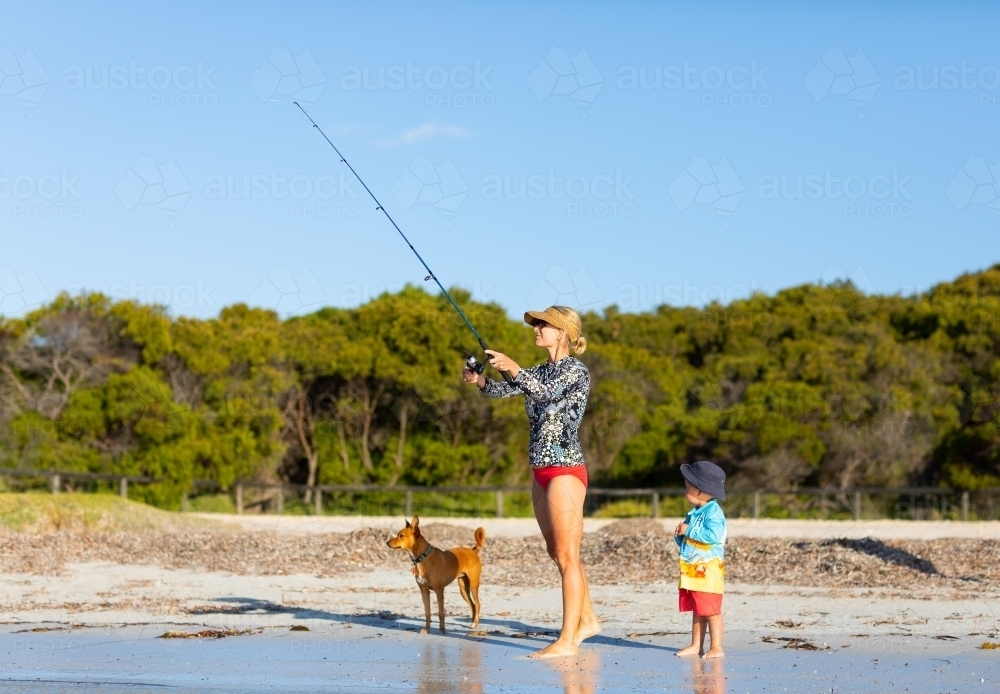 Lady casting fishing rod while toddler and pet dog look on - Australian Stock Image