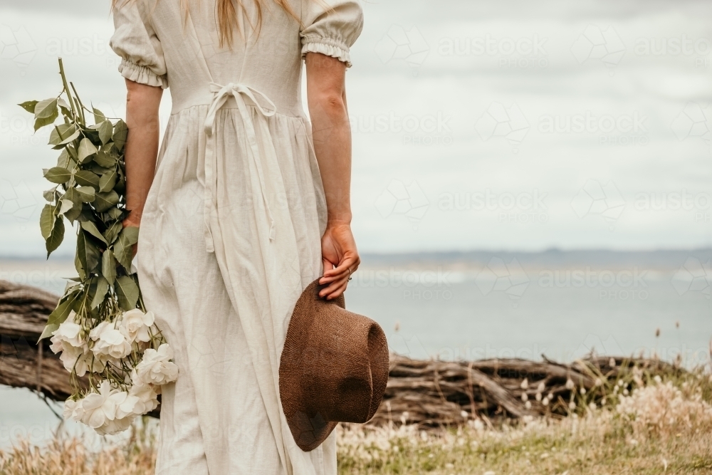 Lady holds flowers and a hat by the sea. - Australian Stock Image