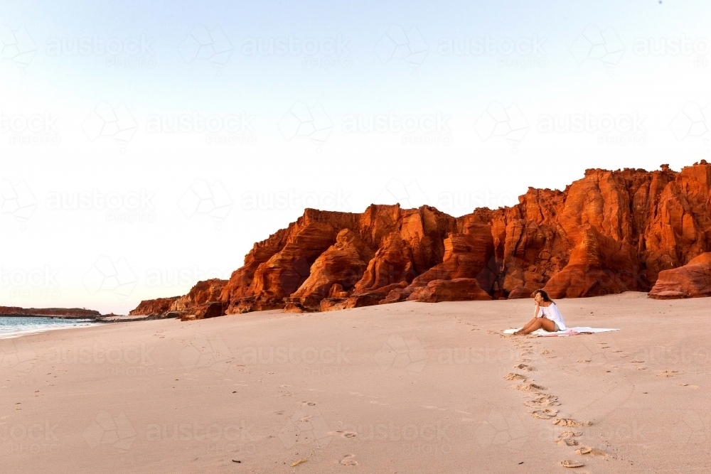 Landscape shot of a woman sitting on the sand with a tall red rock formation beside her - Australian Stock Image
