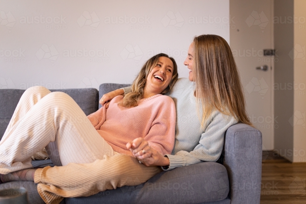 Laughing female same sex couple  in the living room, cuddling on the grey sofa - Australian Stock Image