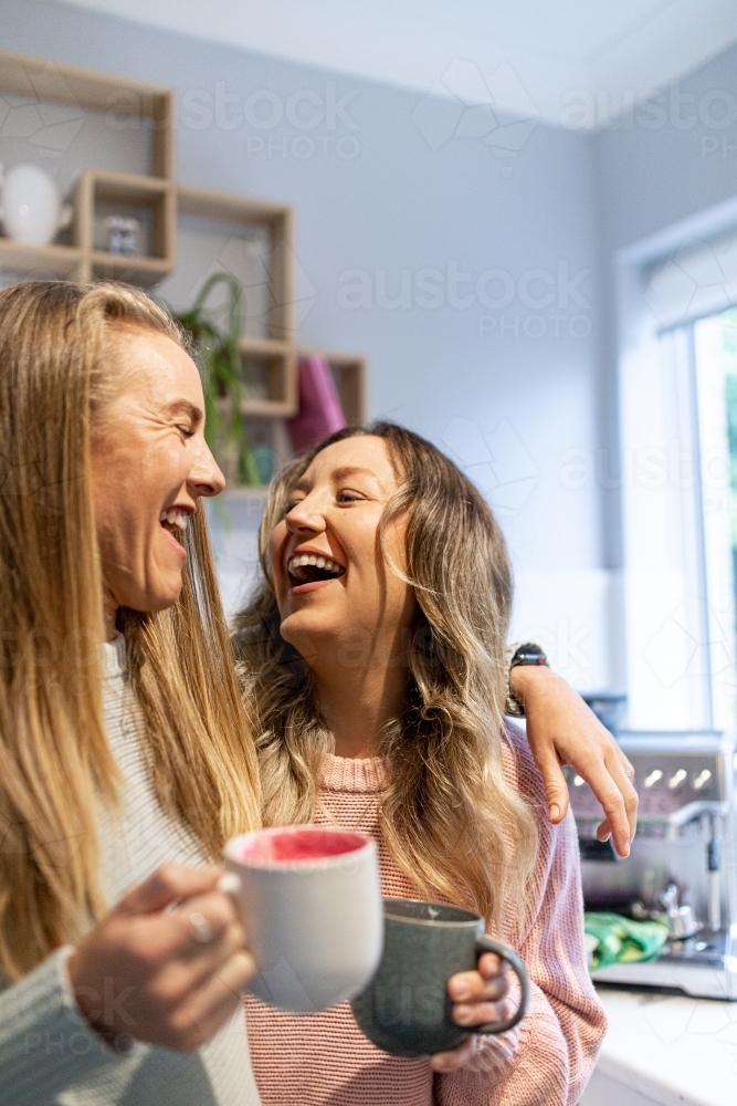 Laughing female same sex couple standing together with coffees in their kitchen - Australian Stock Image