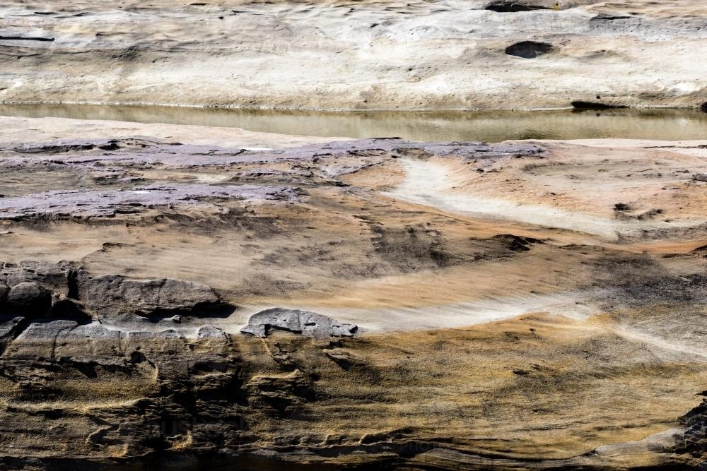 Layered shot of rough patterned rocks and water with shades of purple and orange - Australian Stock Image