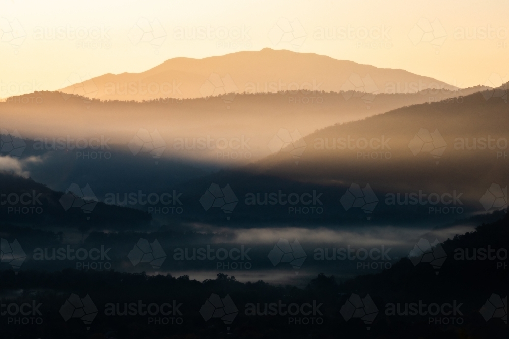 Layers of mountains on a cold foggy morning in winter with the early morning sun shining through - Australian Stock Image