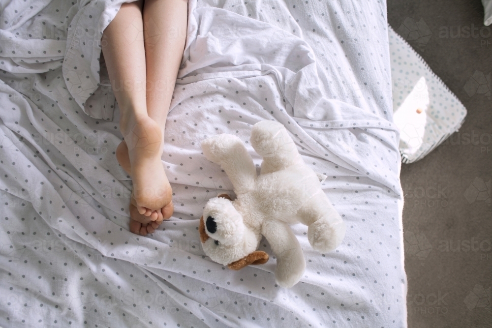 Legs and feet of a child laying in a messy bed with a teddy - Australian Stock Image