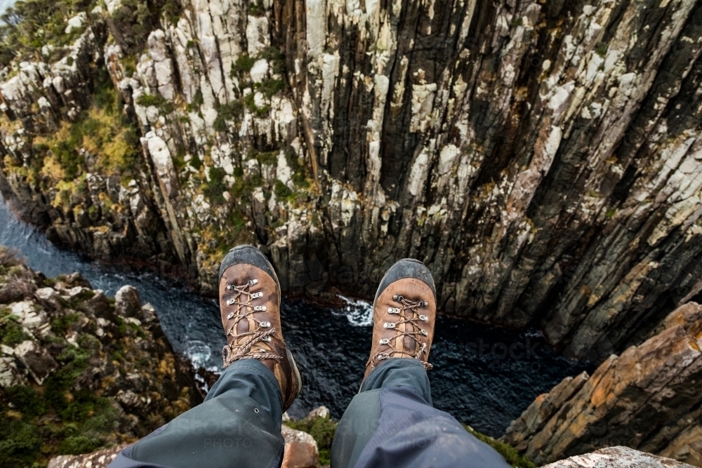 legs and leather boots hanging over a rocky cliff above water - Australian Stock Image