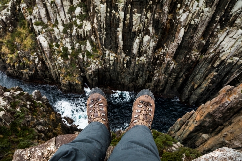 legs and leather boots hanging over a rocky cliff above water - Australian Stock Image