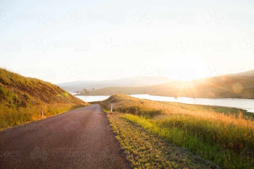 Lens flare and sunlight shining over lake beside road - Australian Stock Image