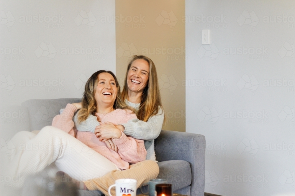 Lesbian couple laughing and hugging on the couch - Australian Stock Image