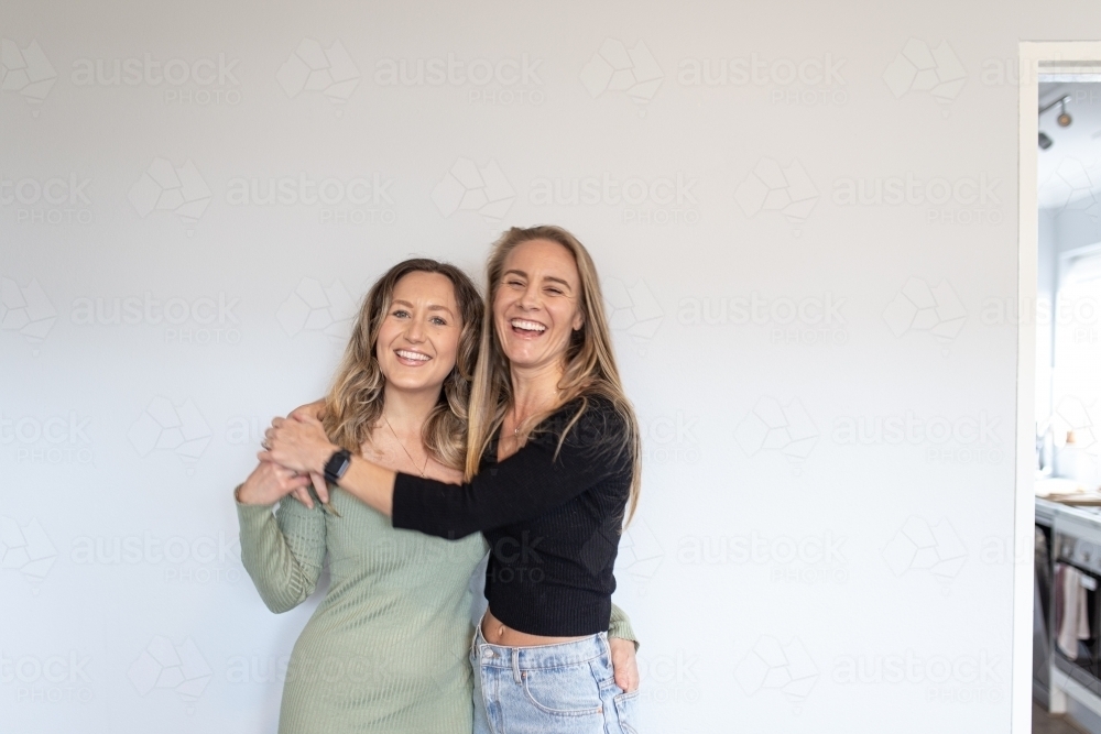 Lesbian couple  leaning against white wall while hugging and wrapping arms around one another - Australian Stock Image