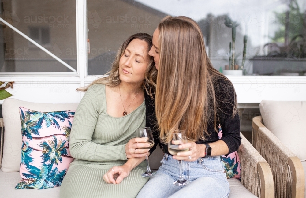 Lesbian couple on outdoor sofa, drinking wine and holding one another - Australian Stock Image