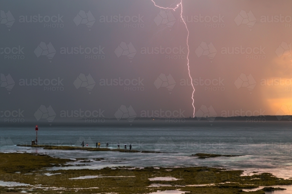 Image of Lightning strikes as rain falls during thunderstorms over fishermen on a rocky outcrop