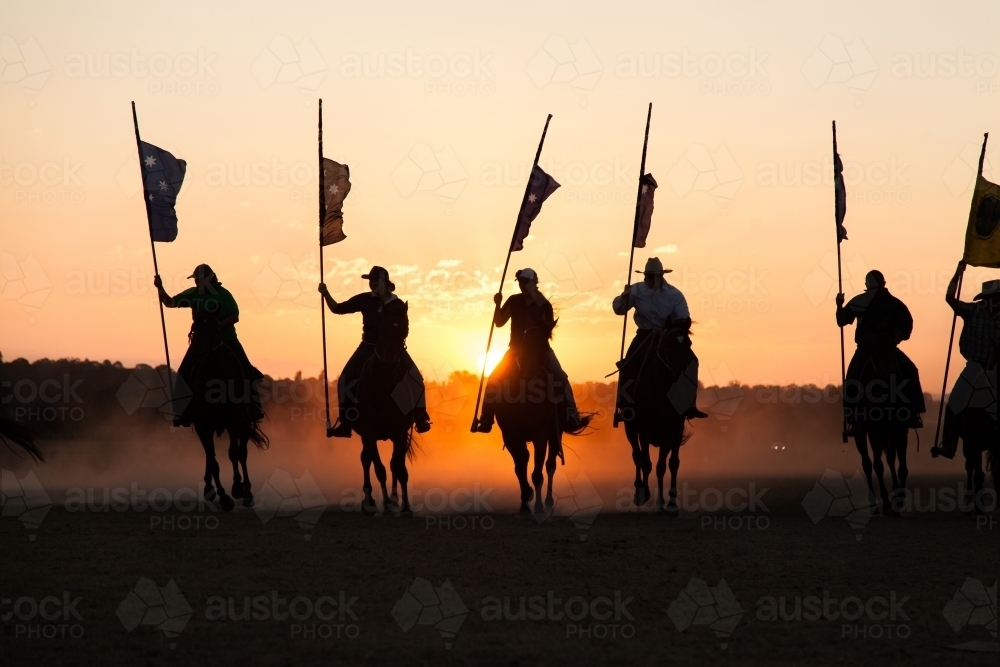 Line of silhouetted horses and riders with flag poles charging at sunset - Australian Stock Image