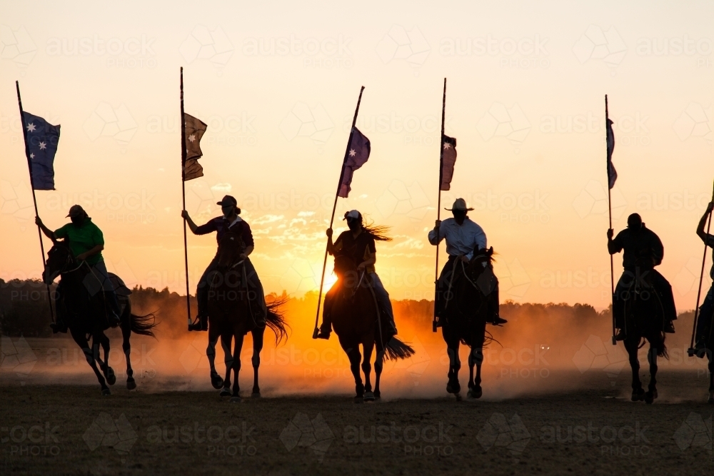 Line of silhouetted horses and riders with flag poles charging at sunset - Australian Stock Image