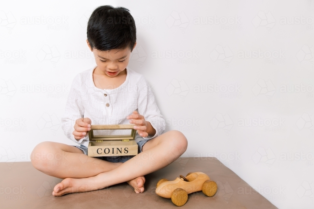 Little asian boy sitting down and putting money into his coin box with his toy car beside him - Australian Stock Image
