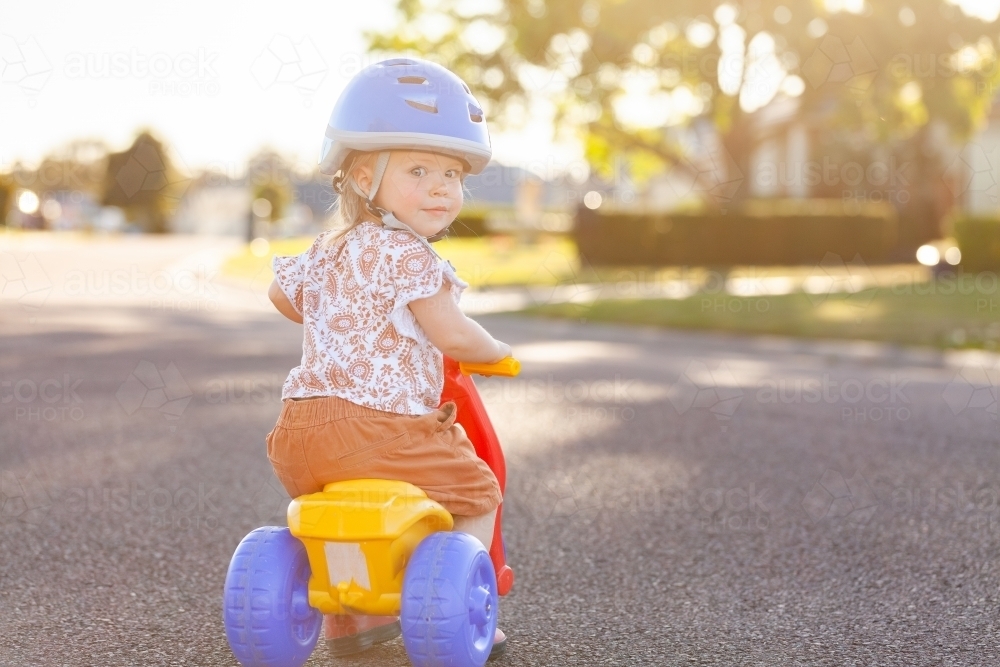 Little Aussie girl riding tricycle down suburban road wearing bike helmet - Australian Stock Image