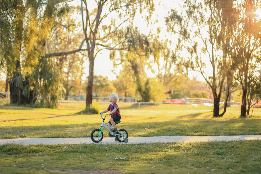 Little blond boy with big happy smile riding bike along walking track in the park - Australian Stock Image