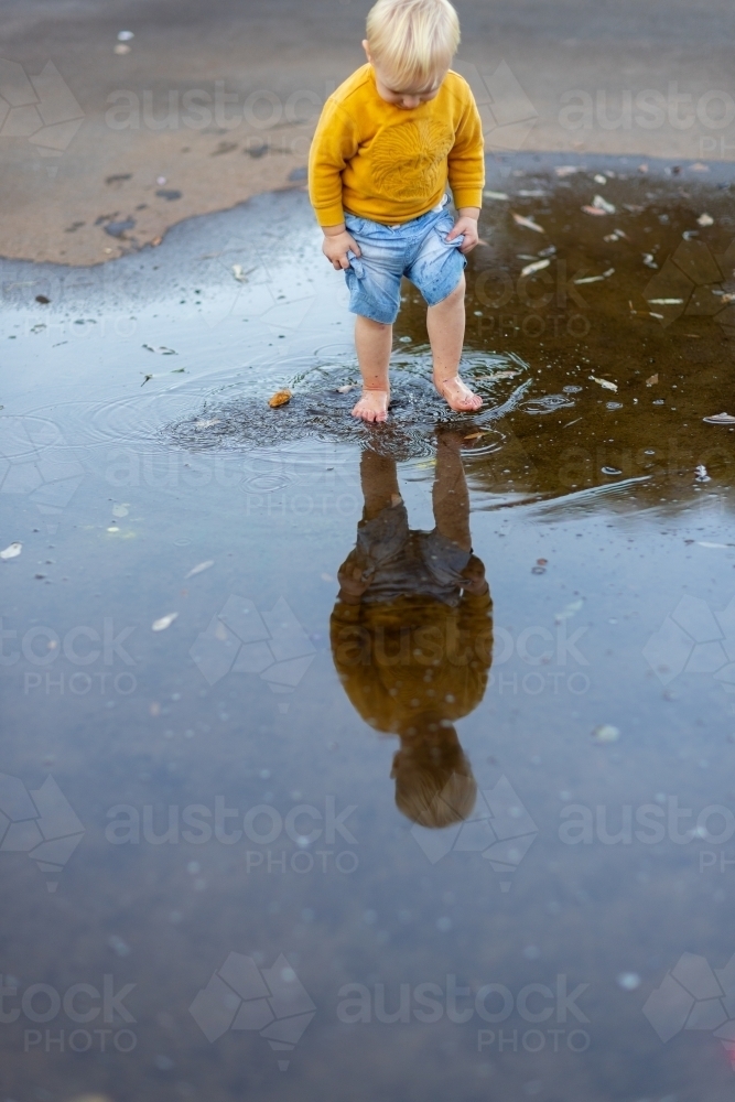 little blonde kid splashing in a puddle with reflection after rain - Australian Stock Image