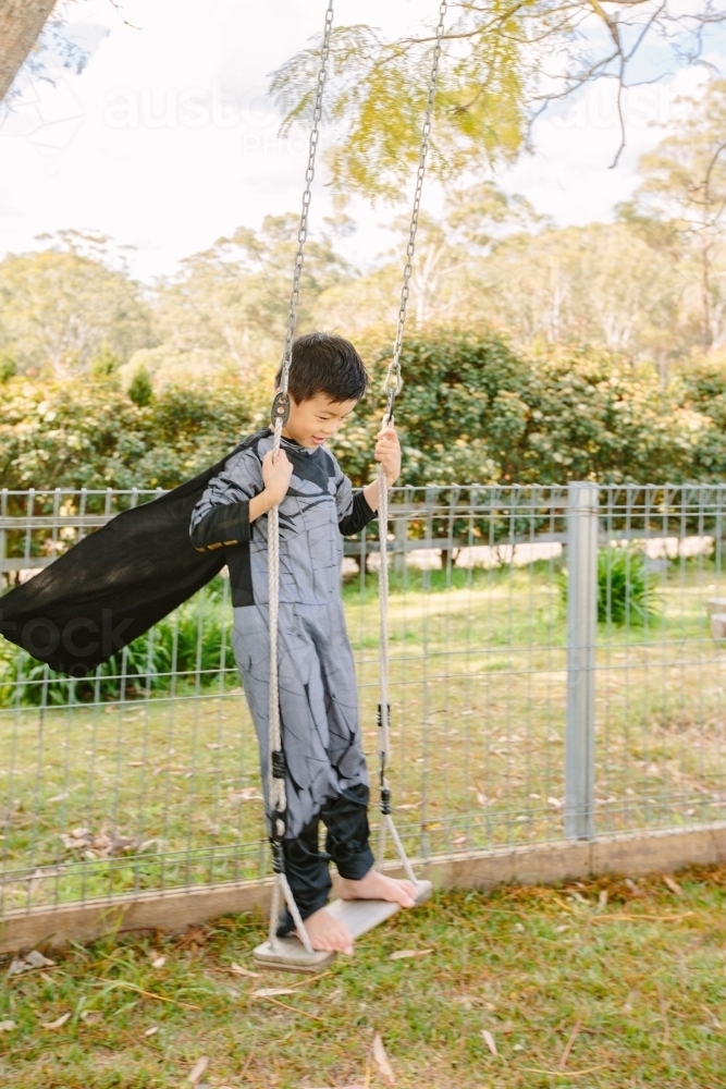 Little boy dressed as batman playing in the garden on a swing on a beautiful day - Australian Stock Image