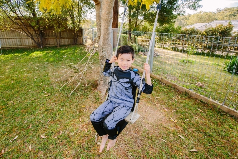 Little boy dressed as batman playing in the garden on a swing on a beautiful day - Australian Stock Image