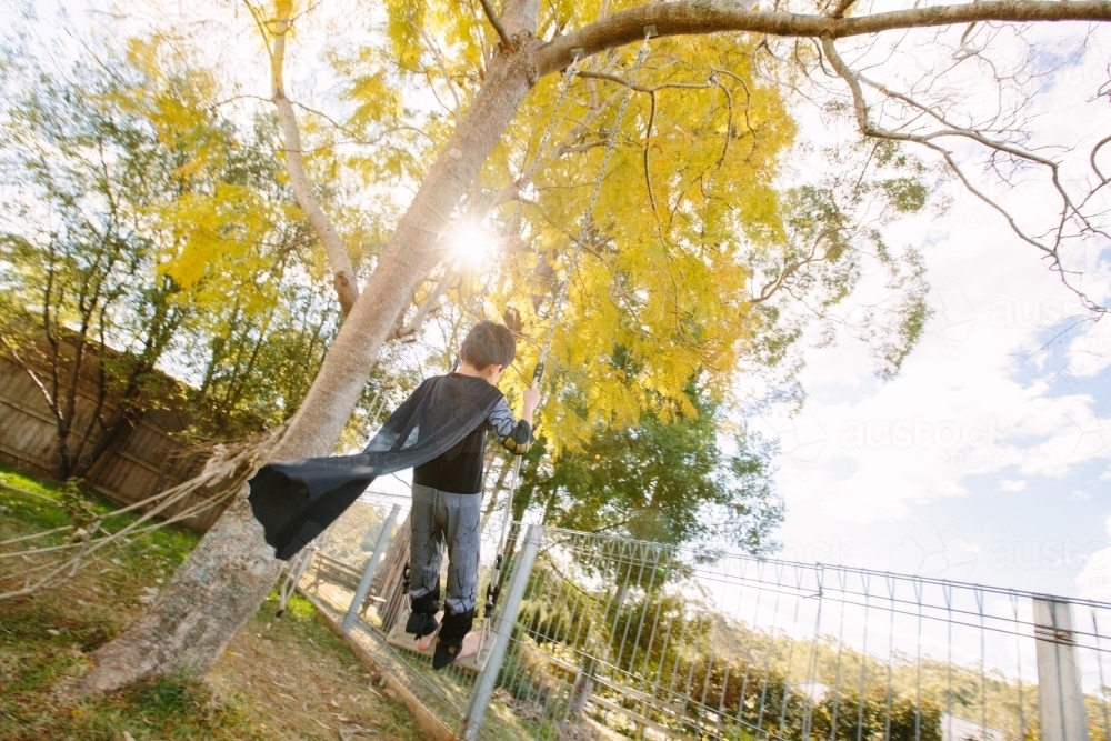 Little boy dressed as batman playing in the garden on a swing on a sunny day - Australian Stock Image