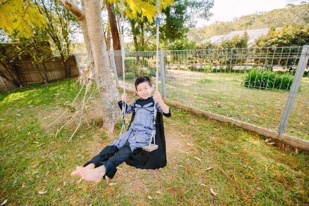 Little boy dressed as batman playing in the garden on a swing on a sunny day - Australian Stock Image