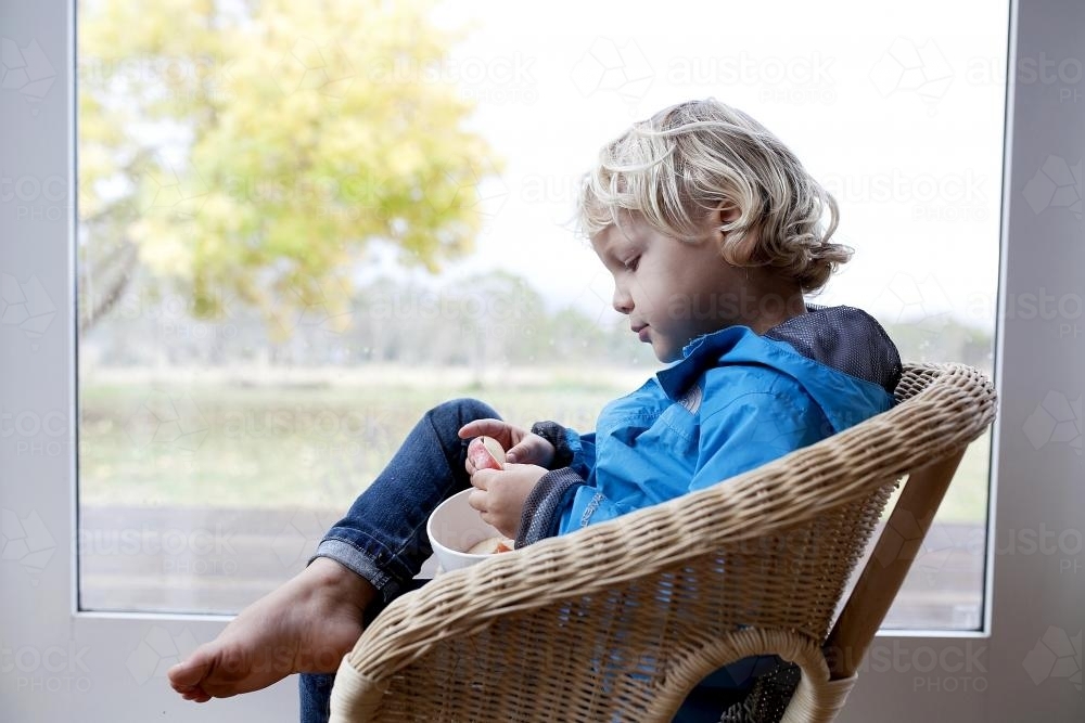 Little boy eating in wicker chair in front of window - Australian Stock Image