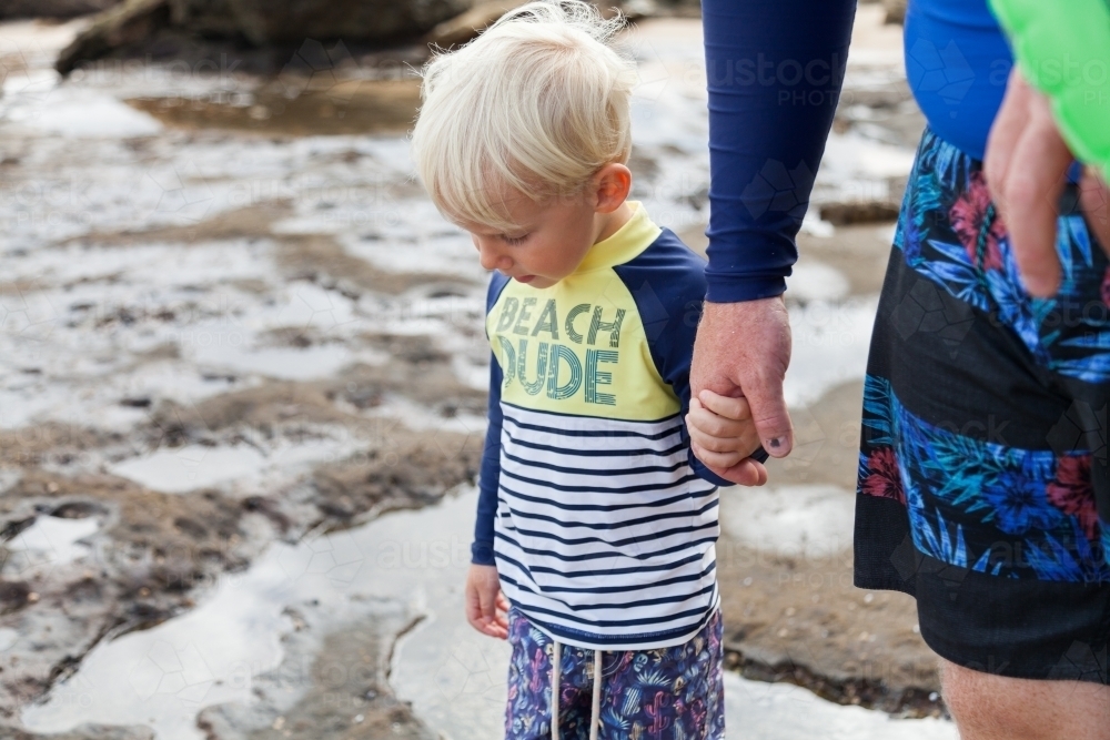 Little boy in swimmers at beach holding fathers hand as they stand in a rock pool - Australian Stock Image