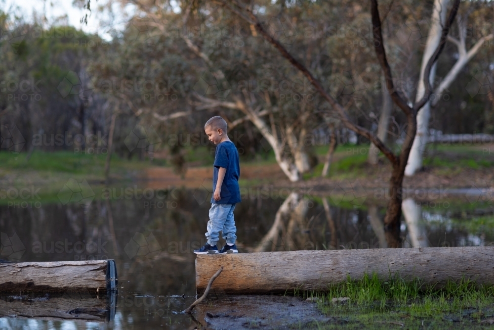 little boy outdoors walking on log at water's edge - Australian Stock Image