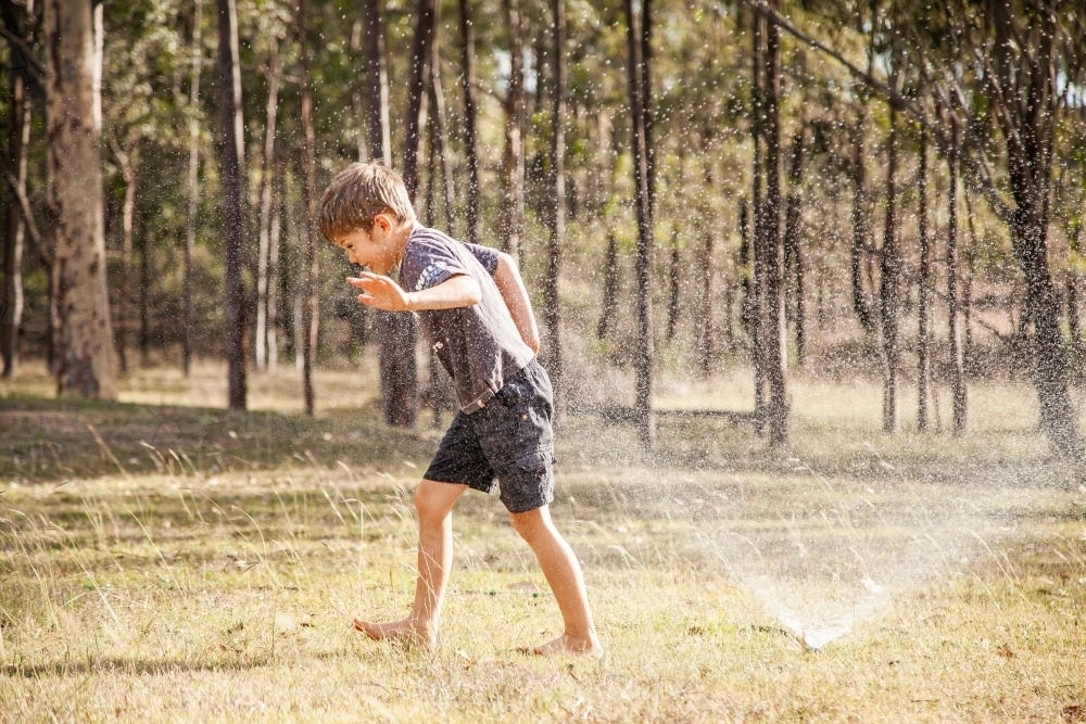 Little boy running under sprinkler in backyard in summer - Australian Stock Image