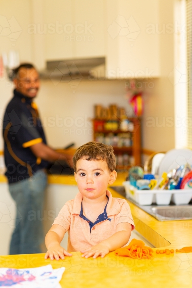 little boy standing at a counter playing with playdough with father in background in kitchen - Australian Stock Image
