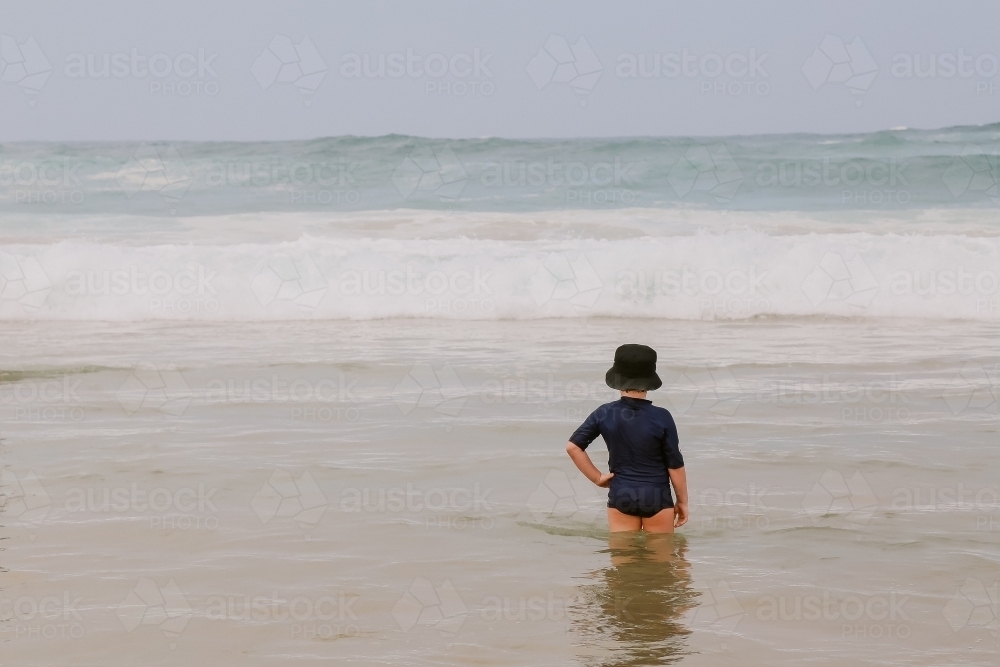 Little boy standing in shallow water at the beach watching waves roll in - Australian Stock Image