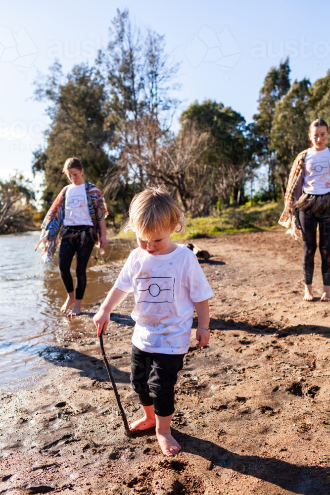 Little boy toddler walking beside river barefoot in mud with stick big sisters following - Australian Stock Image