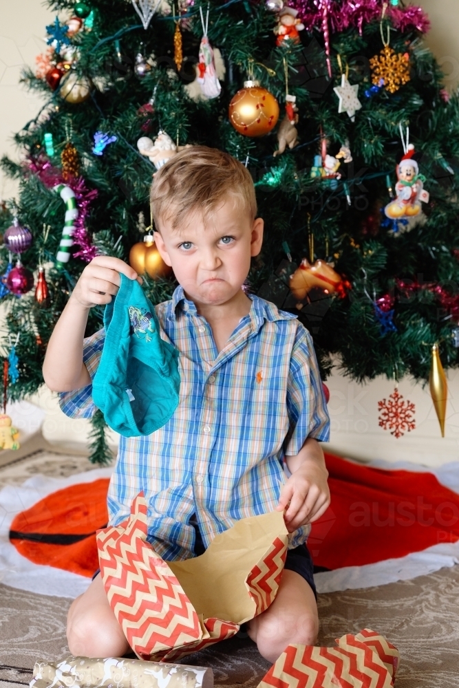 Little boy unhappy with a gift he has opened on Christmas day next to the Christmas tree - Australian Stock Image